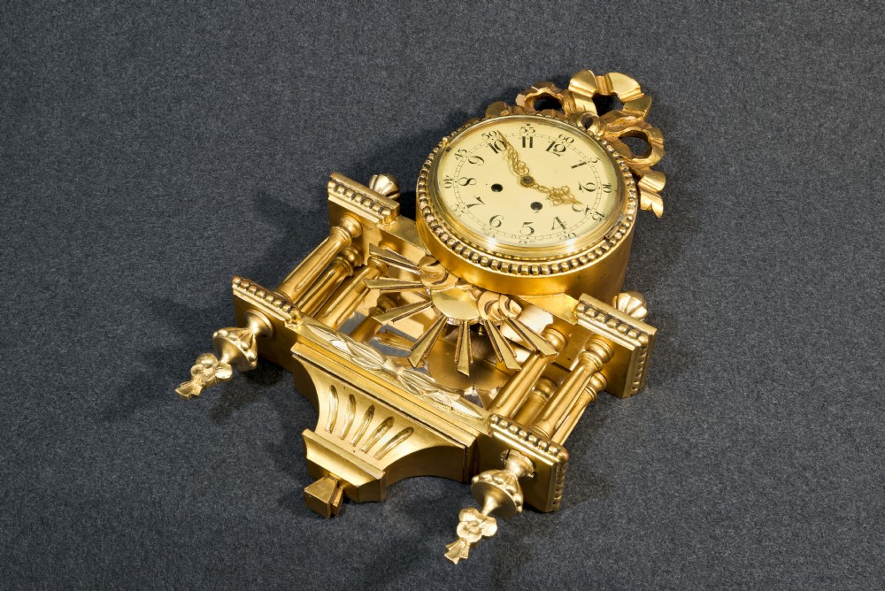 An ornate Swedish Gustavian giltwood cartel wall clock featuring a carved ribbon cresting, twin fluted columns over a mirror recess, and a round dial with Arabic numerals. View IV
