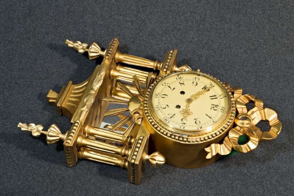 An ornate Swedish Gustavian giltwood cartel wall clock featuring a carved ribbon cresting, twin fluted columns over a mirror recess, and a round dial with Arabic numerals. View II