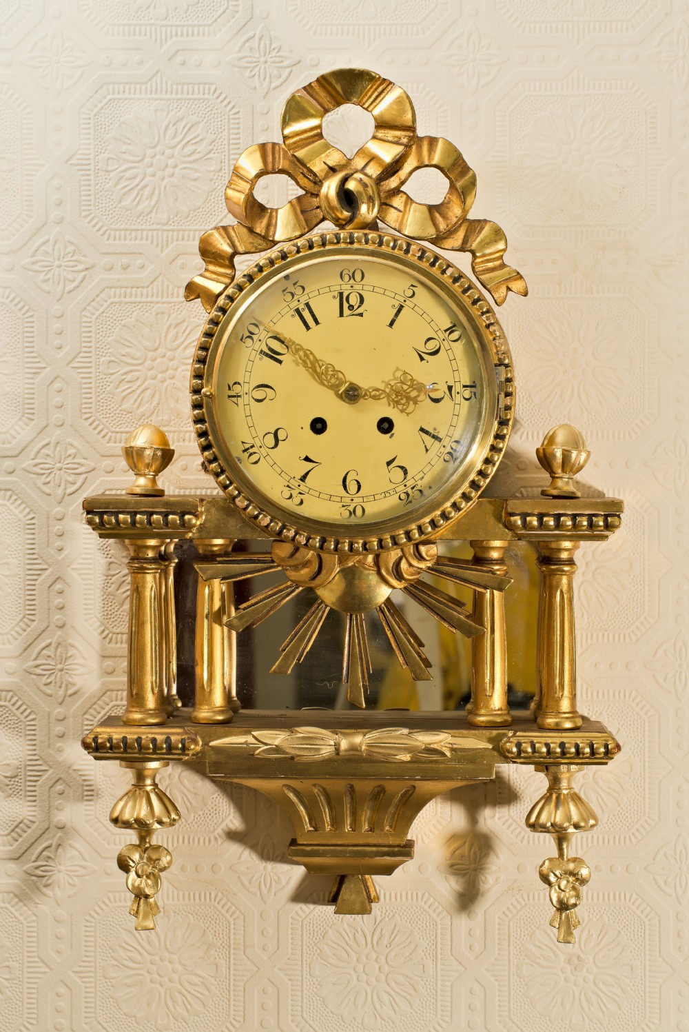 An ornate Swedish Gustavian giltwood cartel wall clock featuring a carved ribbon cresting, twin fluted columns over a mirror recess, and a round dial with Arabic numerals.