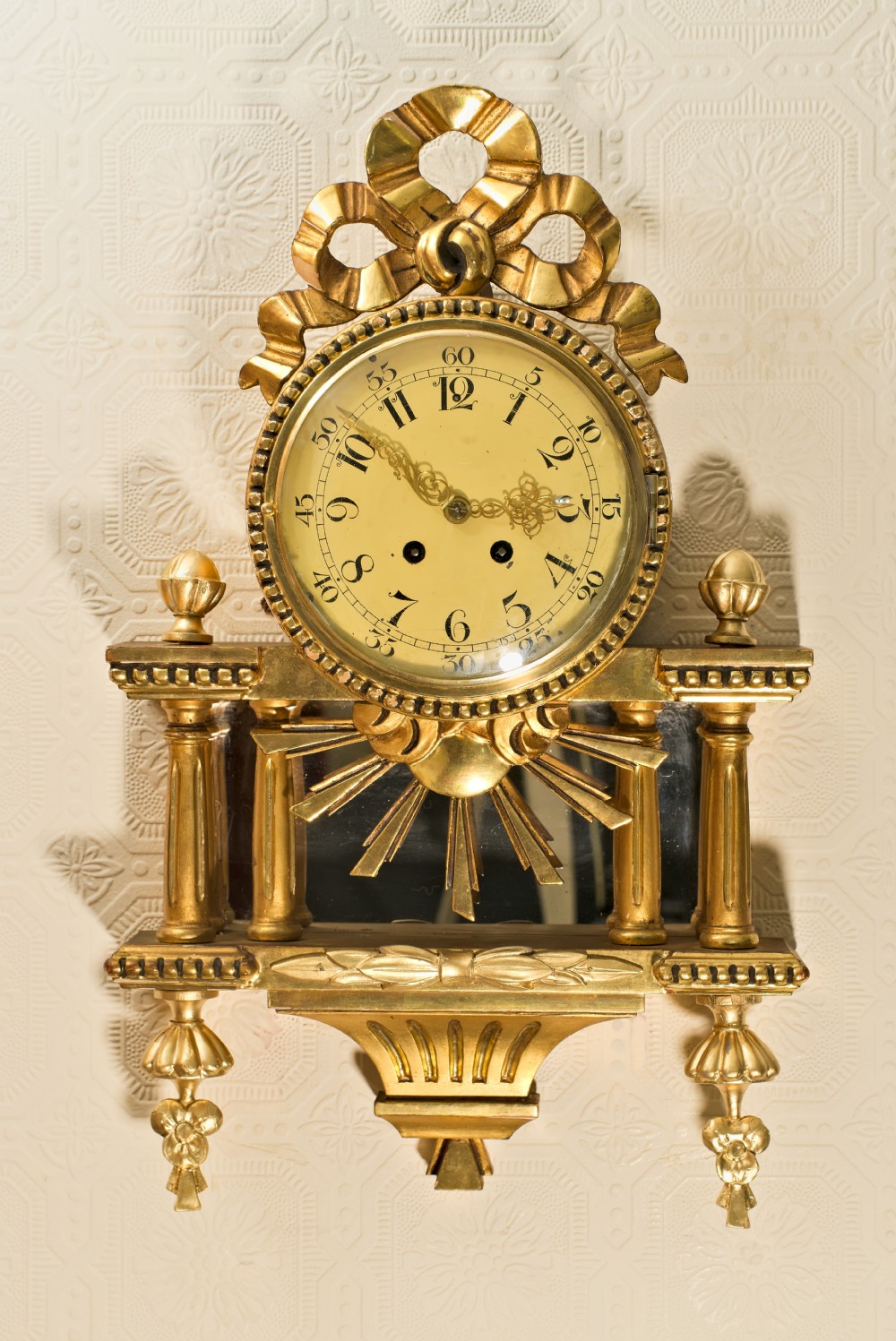 An ornate Swedish Gustavian giltwood cartel wall clock featuring a carved ribbon cresting, twin fluted columns over a mirror recess, and a round dial with Arabic numerals. View V