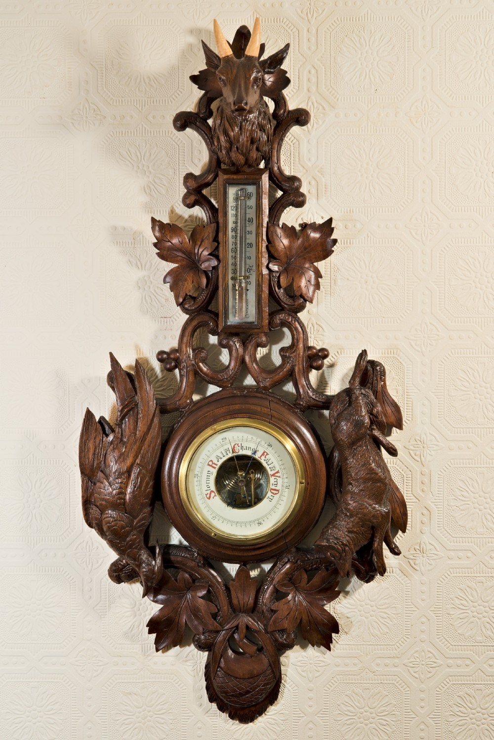 A late 19th-century Black Forest carved wooden wall barometer featuring a chamois head at the top, flanked by carved game animals and foliage around a central dial and upper thermometer tube.