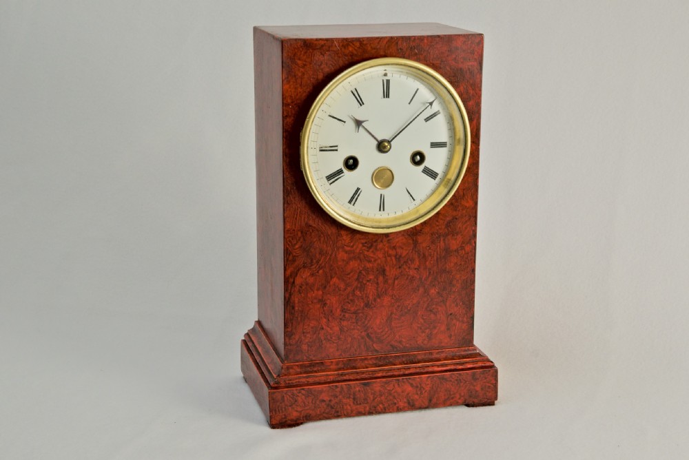 An upright rectangular clock featuring a mottled red faux tortoiseshell case, a stepped base, and a circular white enamel dial framed by a brass bezel.