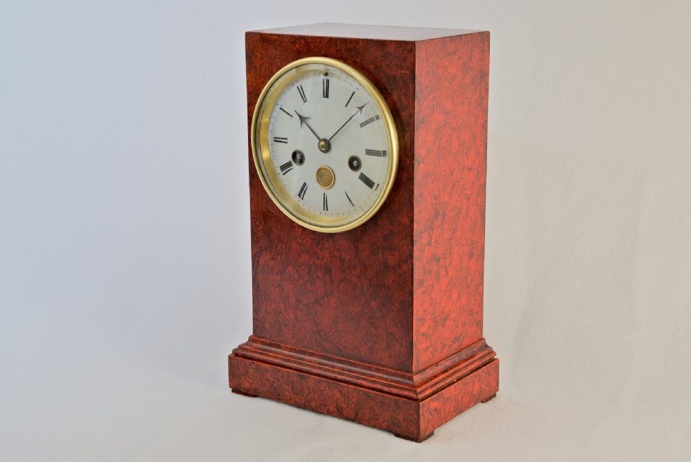 An upright rectangular clock featuring a mottled red faux tortoiseshell case, a stepped base, and a circular white enamel dial framed by a brass bezel. View II