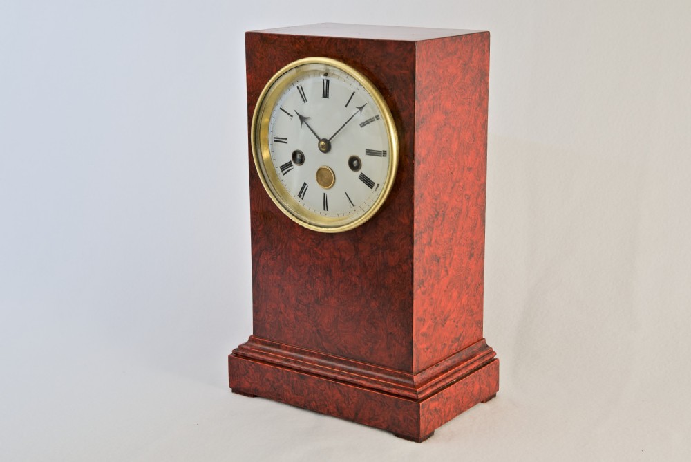 An upright rectangular clock featuring a mottled red faux tortoiseshell case, a stepped base, and a circular white enamel dial framed by a brass bezel. View III