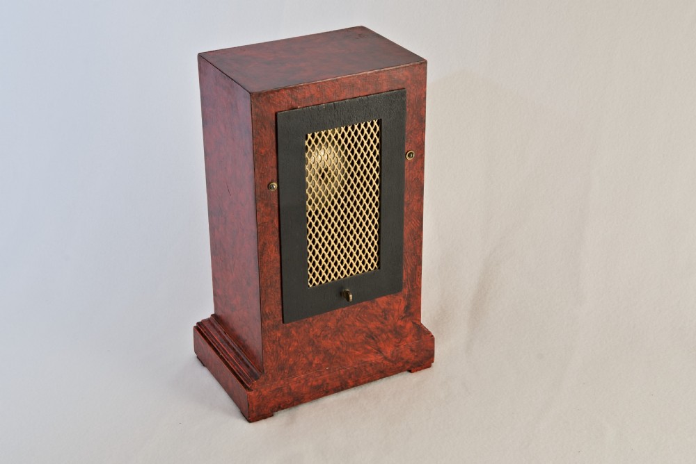 An upright rectangular clock featuring a mottled red faux tortoiseshell case, a stepped base, and a circular white enamel dial framed by a brass bezel. View V