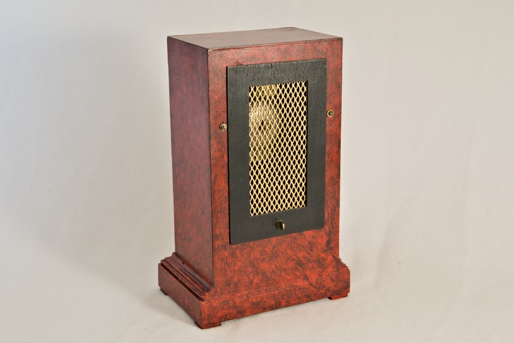 An upright rectangular clock featuring a mottled red faux tortoiseshell case, a stepped base, and a circular white enamel dial framed by a brass bezel. View VI