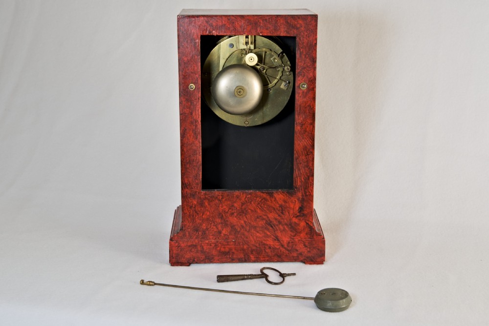 An upright rectangular clock featuring a mottled red faux tortoiseshell case, a stepped base, and a circular white enamel dial framed by a brass bezel. View VII