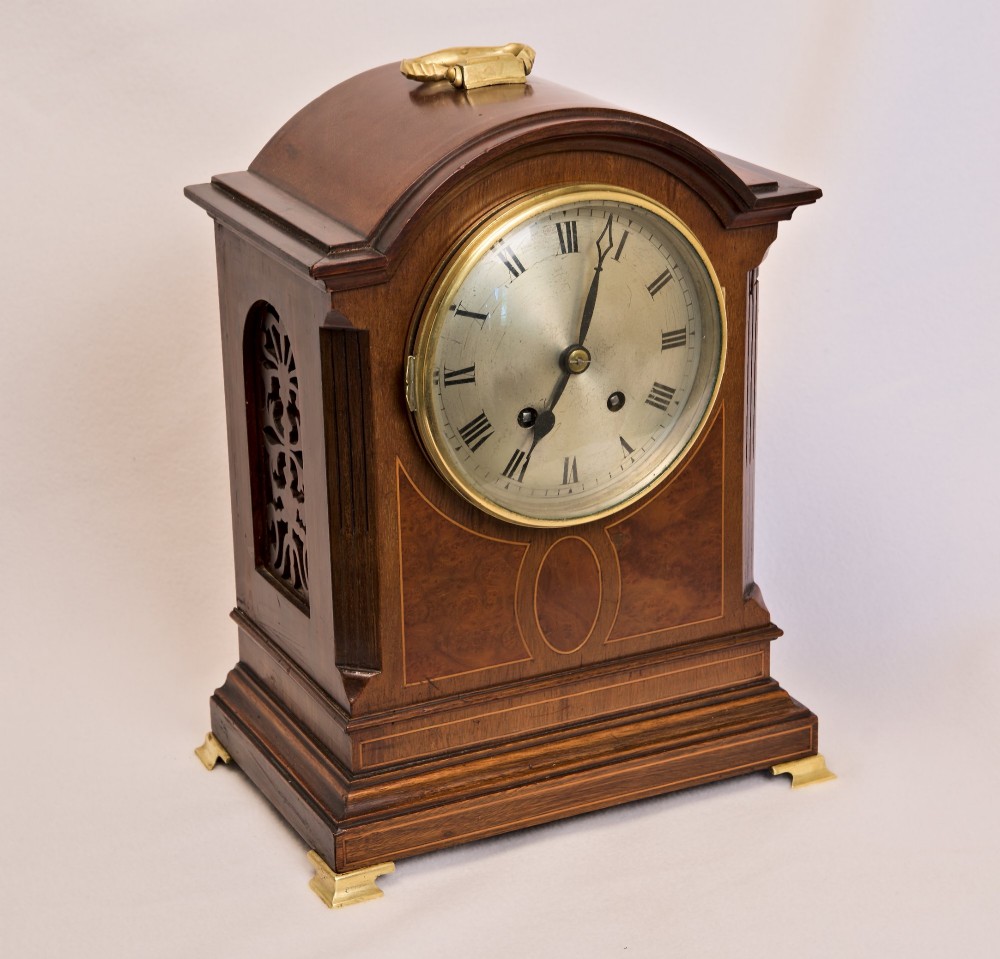 An Edwardian inlaid mahogany arch-top mantel clock with a brass-bezel dial, Roman numerals, side sound frets, and brass corner feet.