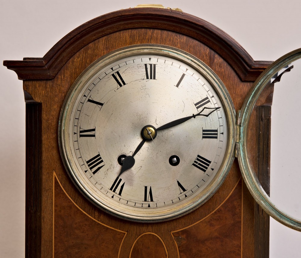 An Edwardian inlaid mahogany arch-top mantel clock with a brass-bezel dial, Roman numerals, side sound frets, and brass corner feet. View IX