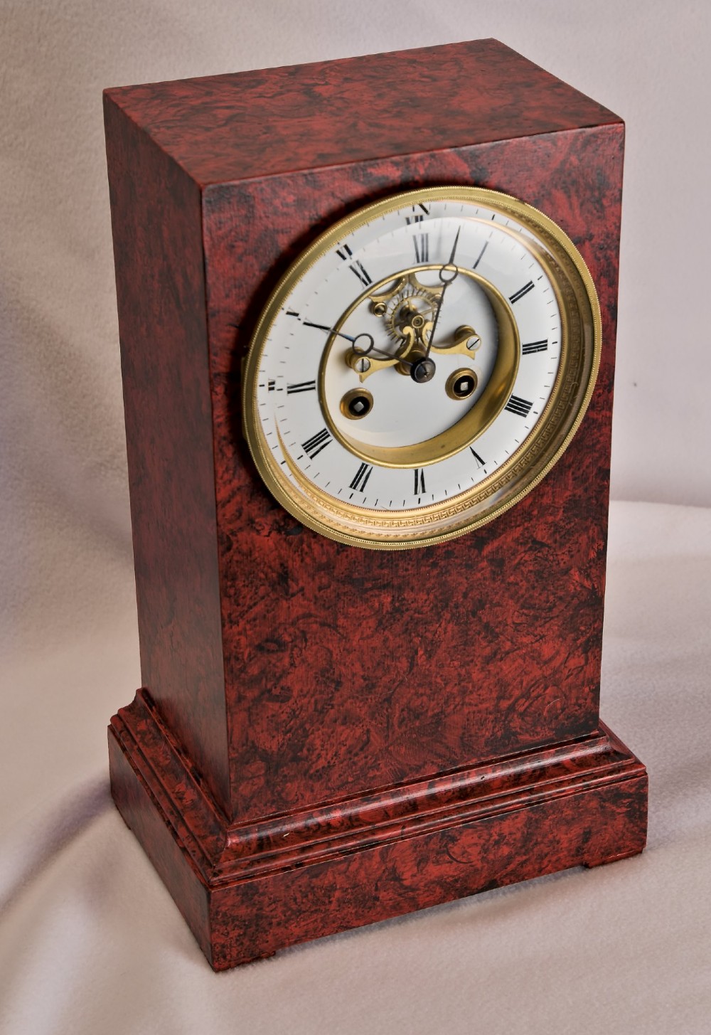 A rectangular mantel clock featuring a red and black faux tortoiseshell finish, a circular brass-framed dial with Roman numerals, and a visible central movement mechanism.