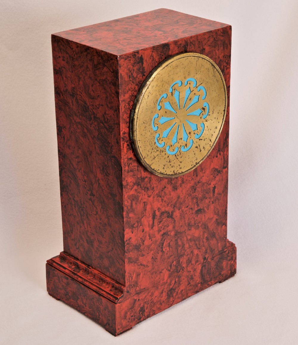 A rectangular mantel clock featuring a red and black faux tortoiseshell finish, a circular brass-framed dial with Roman numerals, and a visible central movement mechanism. View IV