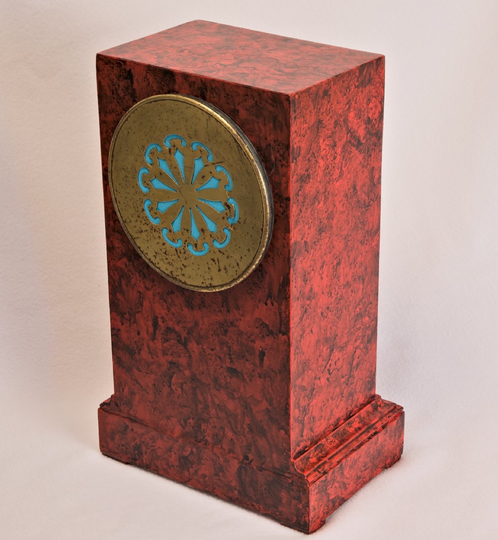 A rectangular mantel clock featuring a red and black faux tortoiseshell finish, a circular brass-framed dial with Roman numerals, and a visible central movement mechanism. View V