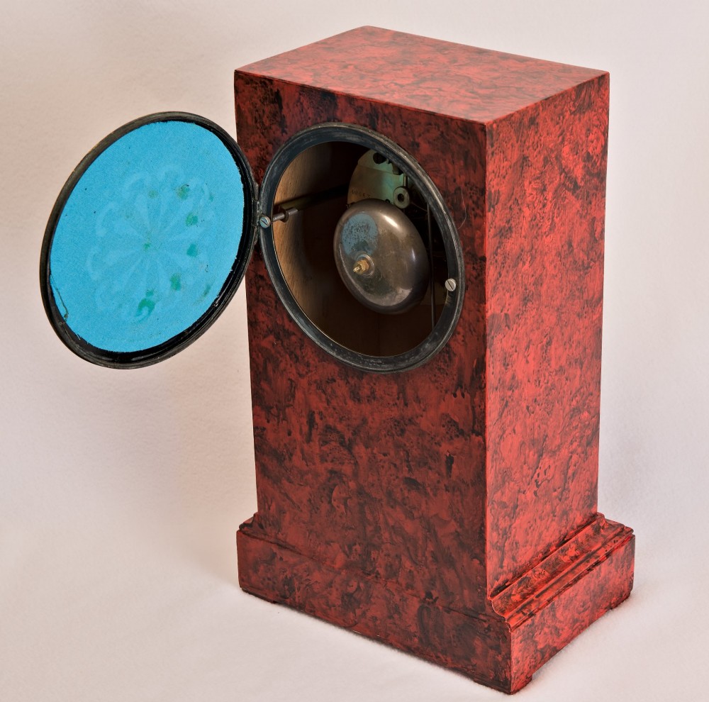 A rectangular mantel clock featuring a red and black faux tortoiseshell finish, a circular brass-framed dial with Roman numerals, and a visible central movement mechanism. View VI