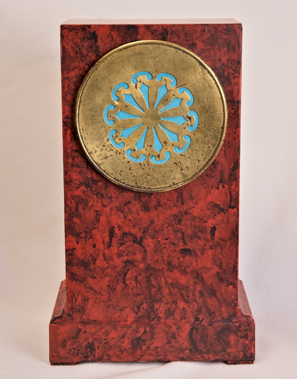 A rectangular mantel clock featuring a red and black faux tortoiseshell finish, a circular brass-framed dial with Roman numerals, and a visible central movement mechanism. View VII