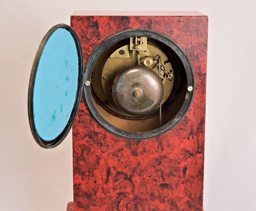 A rectangular mantel clock featuring a red and black faux tortoiseshell finish, a circular brass-framed dial with Roman numerals, and a visible central movement mechanism. View VIII
