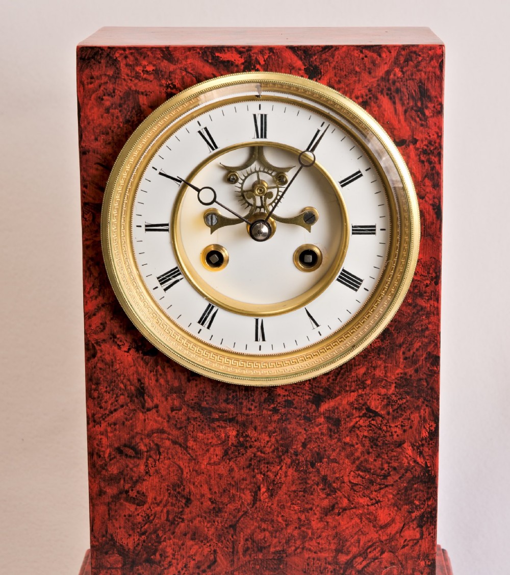 A rectangular mantel clock featuring a red and black faux tortoiseshell finish, a circular brass-framed dial with Roman numerals, and a visible central movement mechanism. View IX