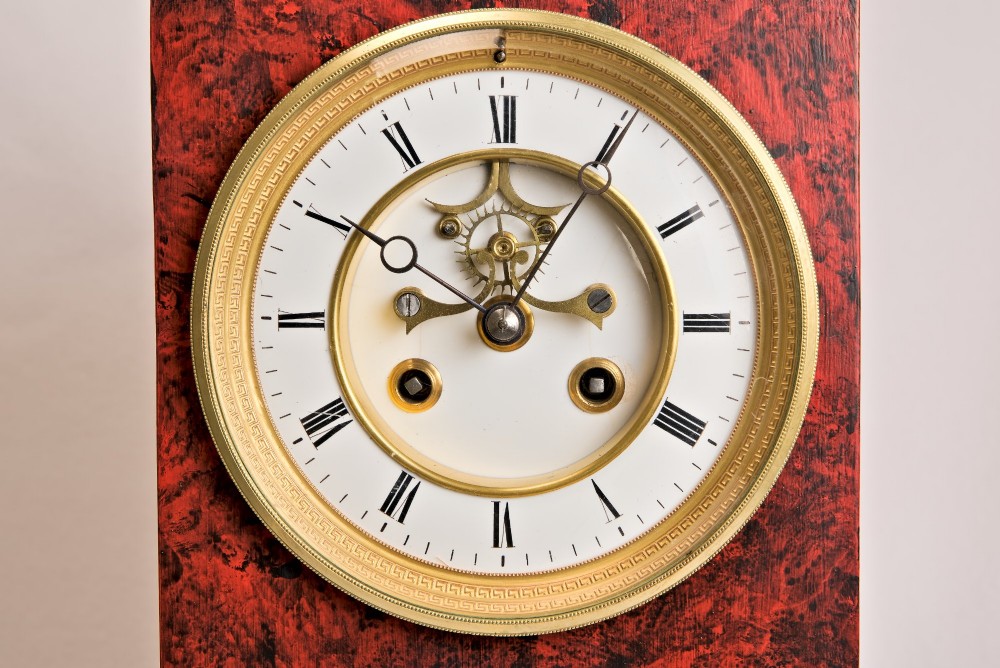 A rectangular mantel clock featuring a red and black faux tortoiseshell finish, a circular brass-framed dial with Roman numerals, and a visible central movement mechanism. View X