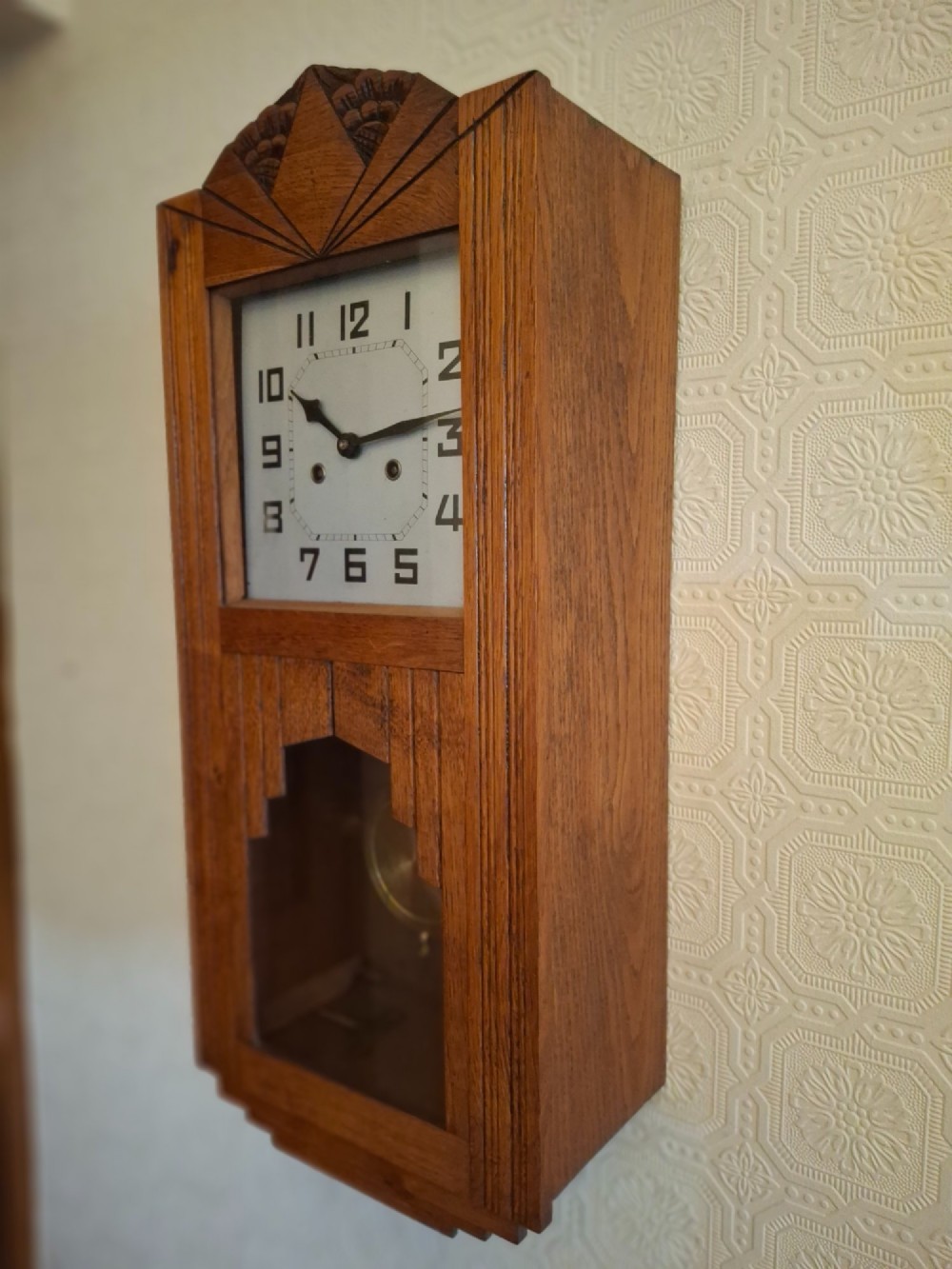 An Art Deco golden oak wall clock featuring a carved sunburst pediment, square face with Arabic numerals, and a lower glass window displaying the pendulum.