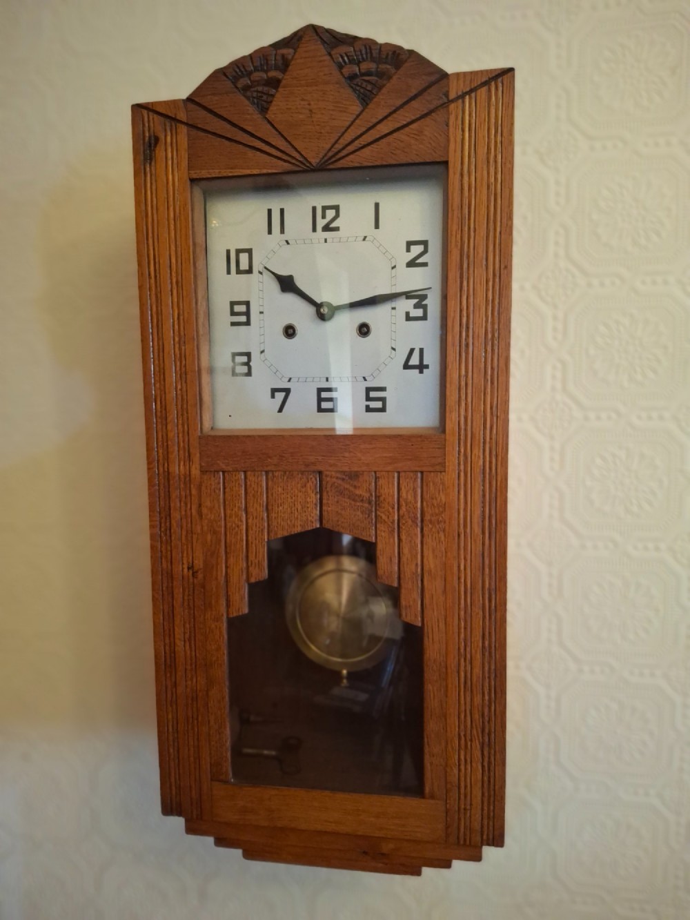 An Art Deco golden oak wall clock featuring a carved sunburst pediment, square face with Arabic numerals, and a lower glass window displaying the pendulum. View II
