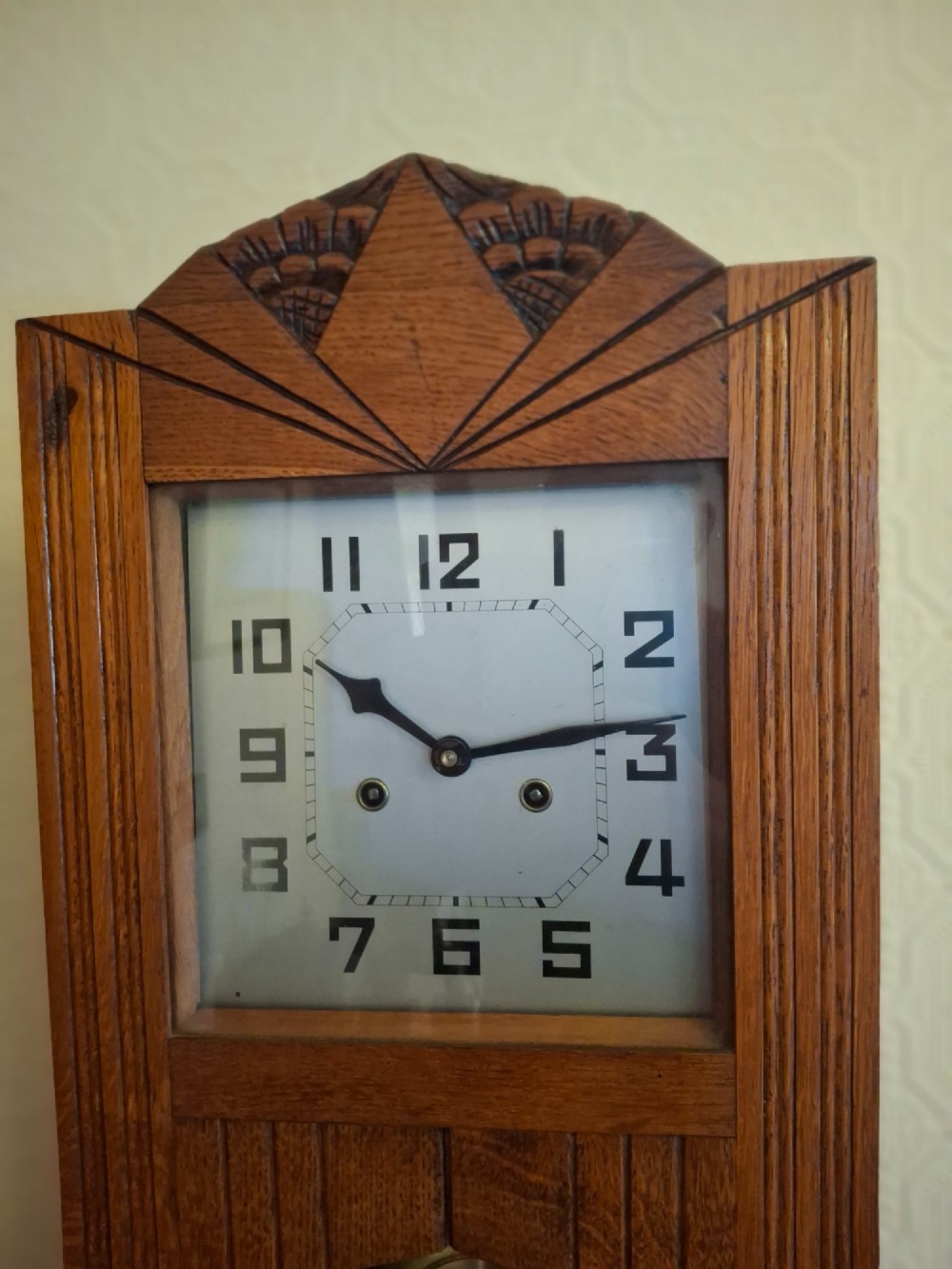 An Art Deco golden oak wall clock featuring a carved sunburst pediment, square face with Arabic numerals, and a lower glass window displaying the pendulum. View III