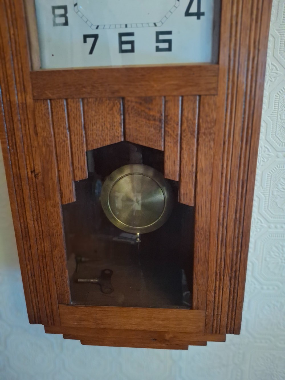 An Art Deco golden oak wall clock featuring a carved sunburst pediment, square face with Arabic numerals, and a lower glass window displaying the pendulum. View IV