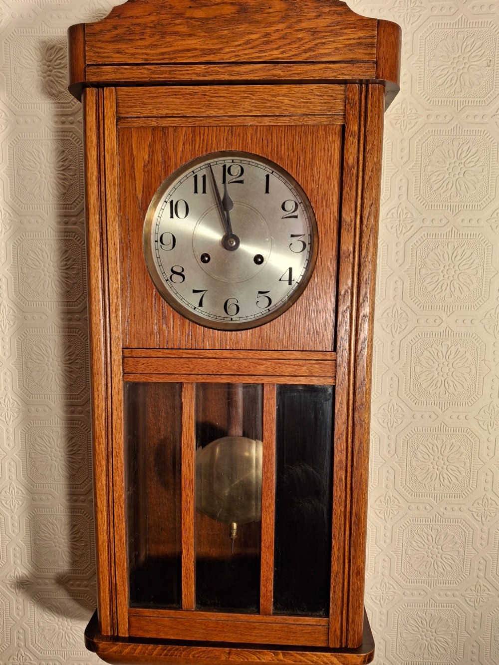 An Arts and Crafts period golden oak wall clock featuring a circular metal dial with Arabic numerals and a lower glazed window displaying a brass pendulum.