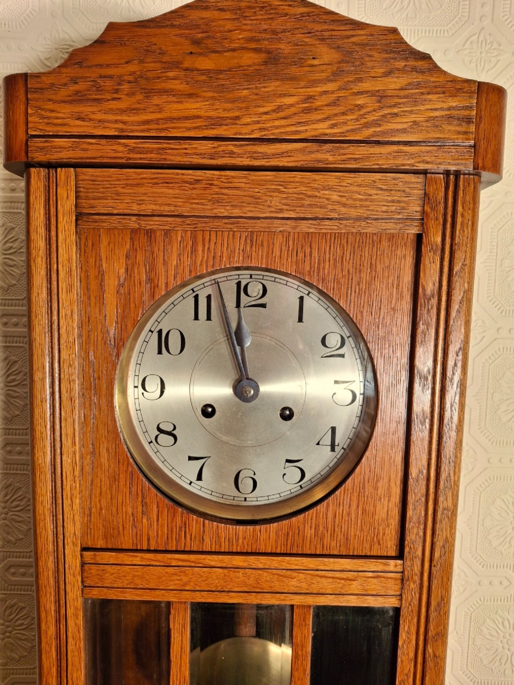 An Arts and Crafts period golden oak wall clock featuring a circular metal dial with Arabic numerals and a lower glazed window displaying a brass pendulum. View III