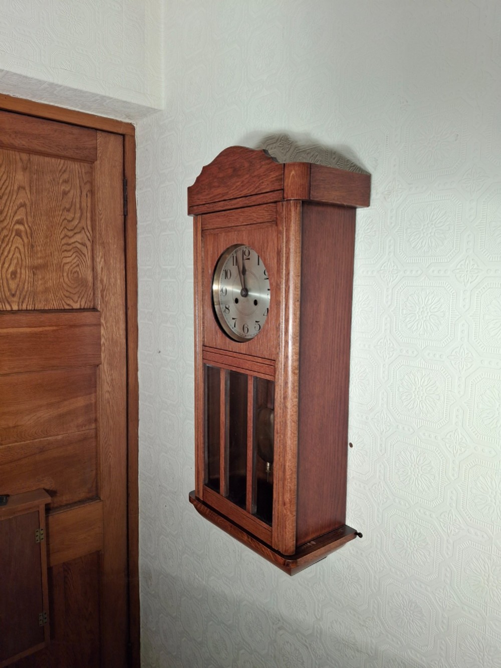 An Arts and Crafts period golden oak wall clock featuring a circular metal dial with Arabic numerals and a lower glazed window displaying a brass pendulum. View IV