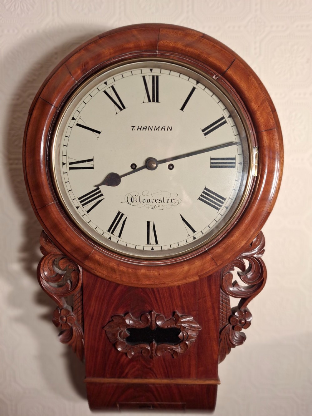 An antique mahogany drop-dial wall clock featuring a circular white dial with Roman numerals above a carved wooden lower trunk.