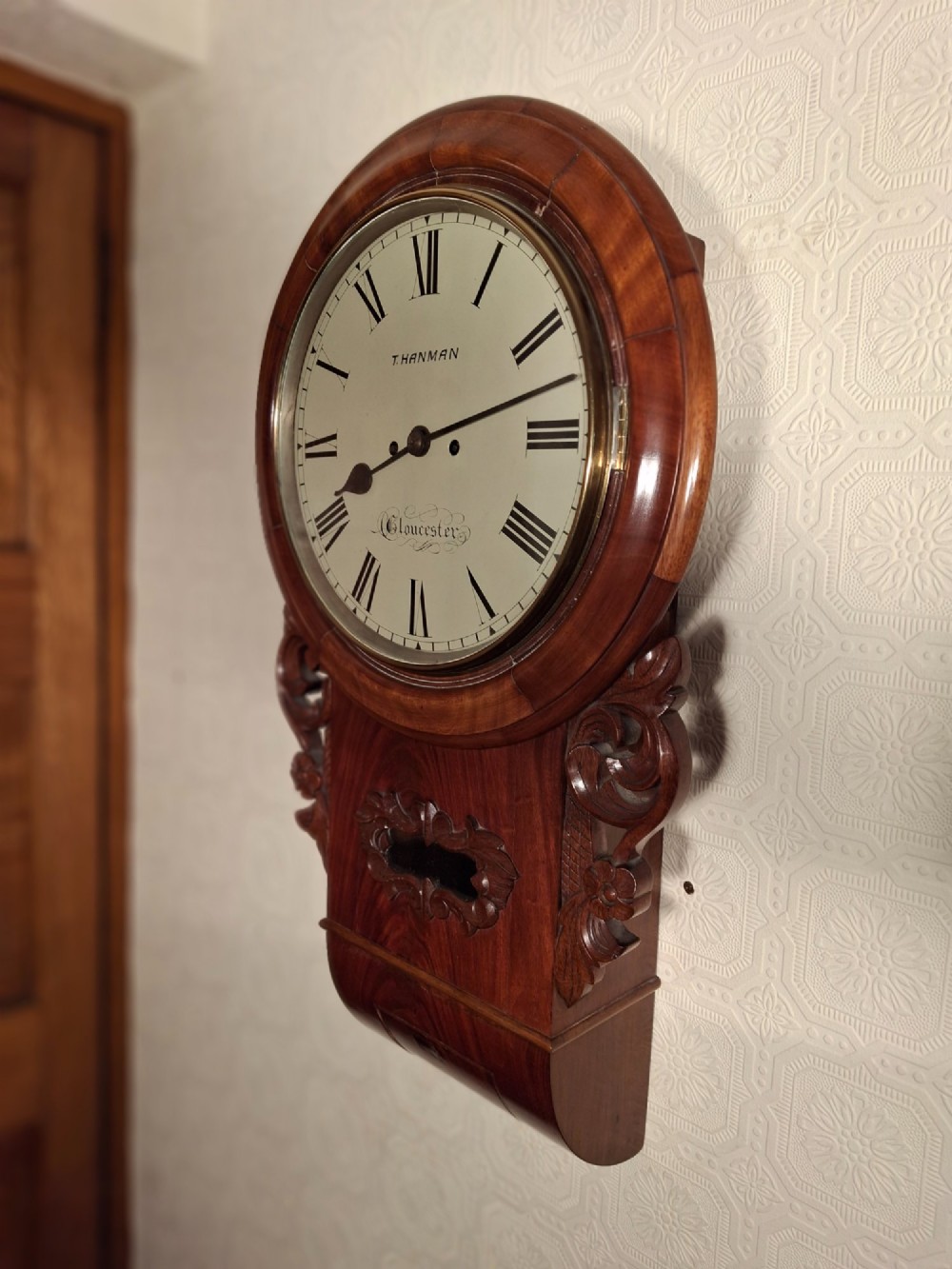 An antique mahogany drop-dial wall clock featuring a circular white dial with Roman numerals above a carved wooden lower trunk. View II