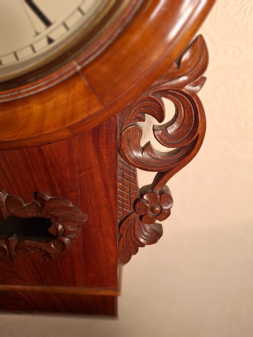 An antique mahogany drop-dial wall clock featuring a circular white dial with Roman numerals above a carved wooden lower trunk. View IV