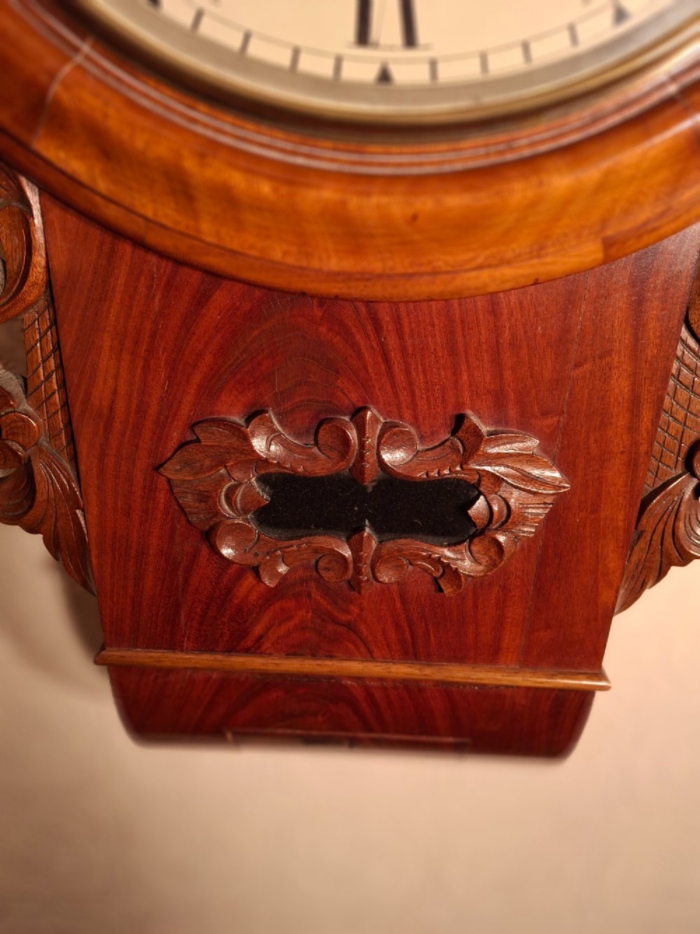 An antique mahogany drop-dial wall clock featuring a circular white dial with Roman numerals above a carved wooden lower trunk. View V
