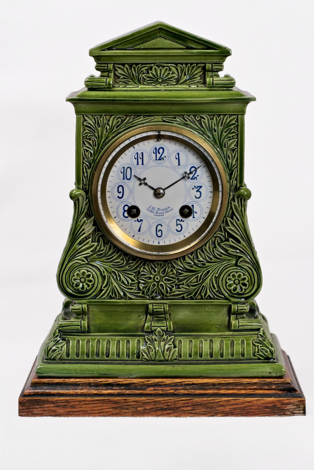 An olive green glazed ceramic mantel clock featuring relief acanthus foliage details, a white dial with blue Arabic numerals, mounted on a dark wooden base.