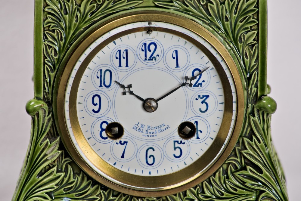 An olive green glazed ceramic mantel clock featuring relief acanthus foliage details, a white dial with blue Arabic numerals, mounted on a dark wooden base. View II