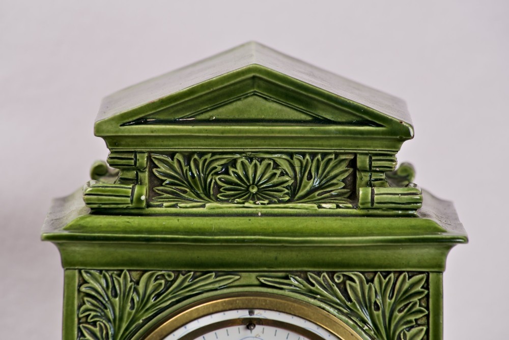An olive green glazed ceramic mantel clock featuring relief acanthus foliage details, a white dial with blue Arabic numerals, mounted on a dark wooden base. View III
