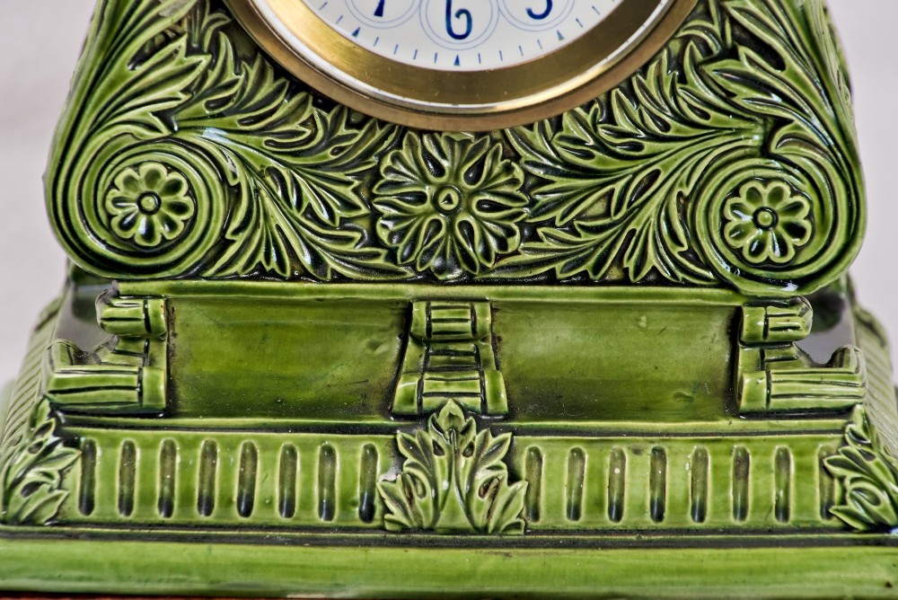An olive green glazed ceramic mantel clock featuring relief acanthus foliage details, a white dial with blue Arabic numerals, mounted on a dark wooden base. View IV