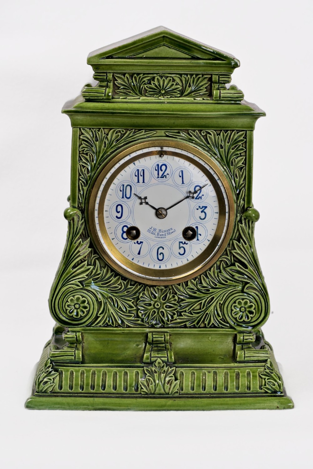 An olive green glazed ceramic mantel clock featuring relief acanthus foliage details, a white dial with blue Arabic numerals, mounted on a dark wooden base. View V