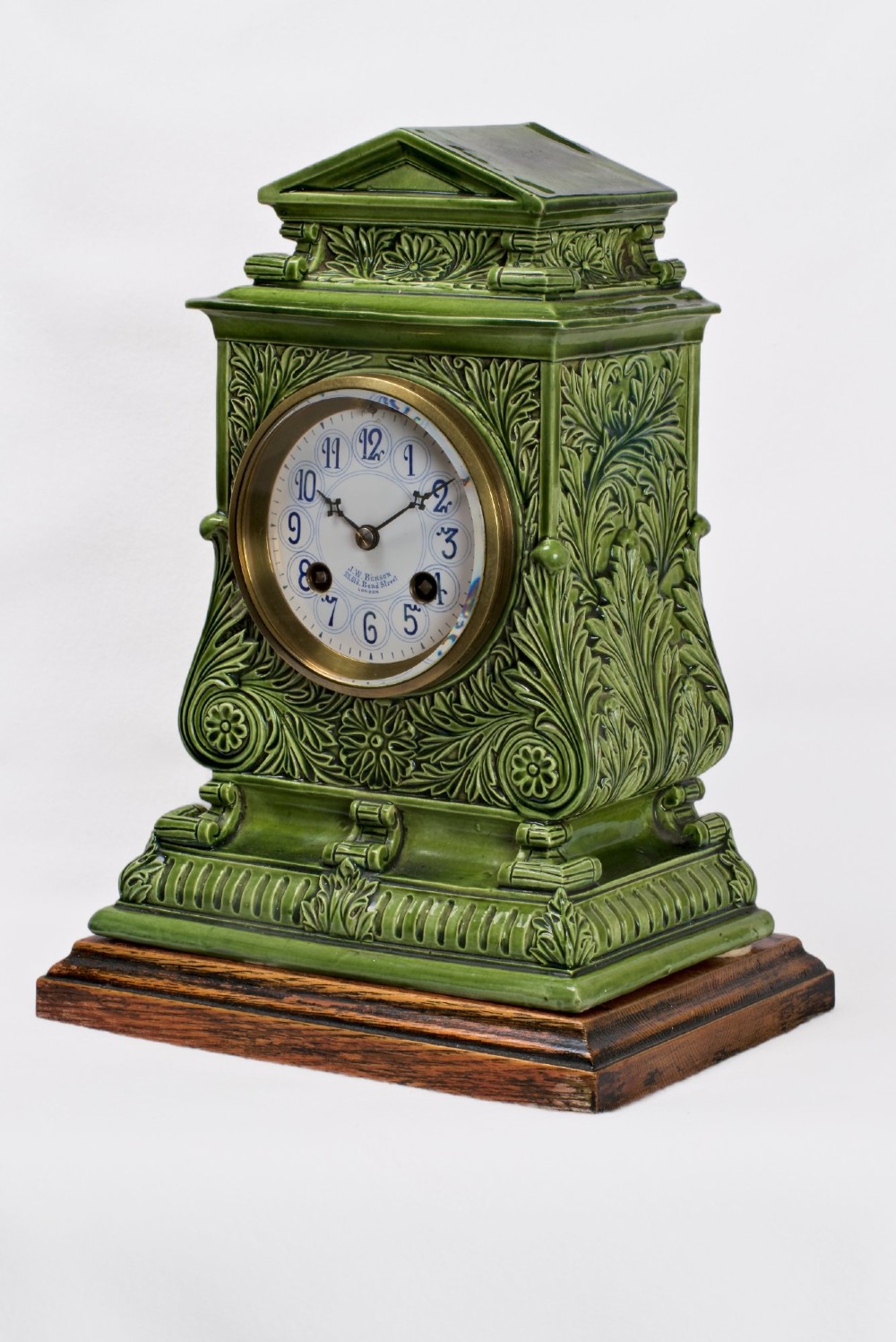 An olive green glazed ceramic mantel clock featuring relief acanthus foliage details, a white dial with blue Arabic numerals, mounted on a dark wooden base. View VI