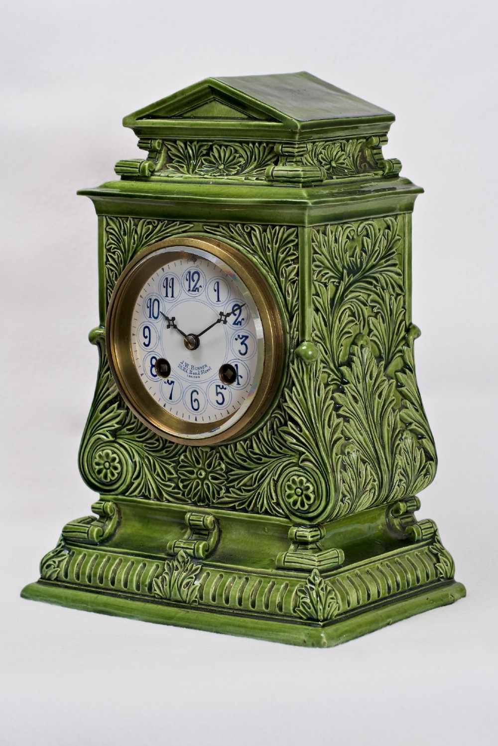 An olive green glazed ceramic mantel clock featuring relief acanthus foliage details, a white dial with blue Arabic numerals, mounted on a dark wooden base. View VII