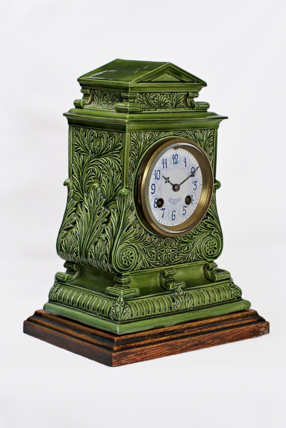 An olive green glazed ceramic mantel clock featuring relief acanthus foliage details, a white dial with blue Arabic numerals, mounted on a dark wooden base. View XI