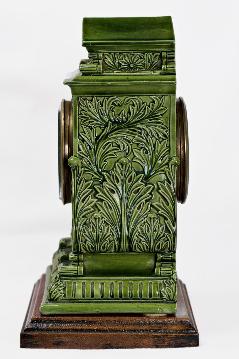 An olive green glazed ceramic mantel clock featuring relief acanthus foliage details, a white dial with blue Arabic numerals, mounted on a dark wooden base. View XII