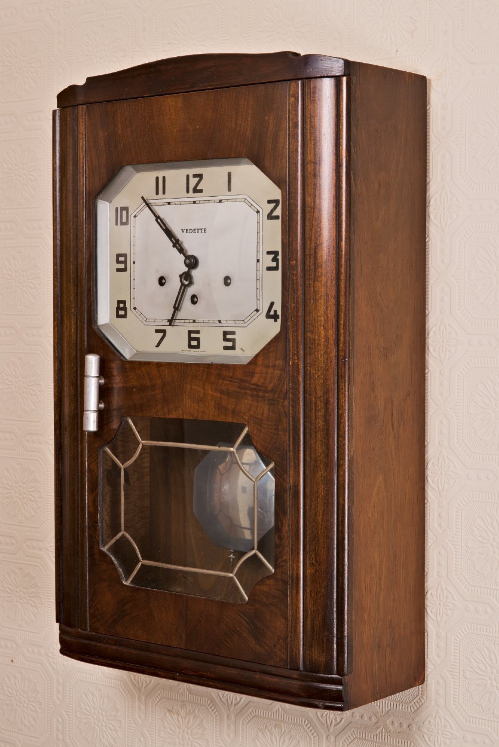 An Art Deco rectangular wooden wall clock featuring a light octagonal dial with Arabic numerals above a geometric leaded glass window. View VI