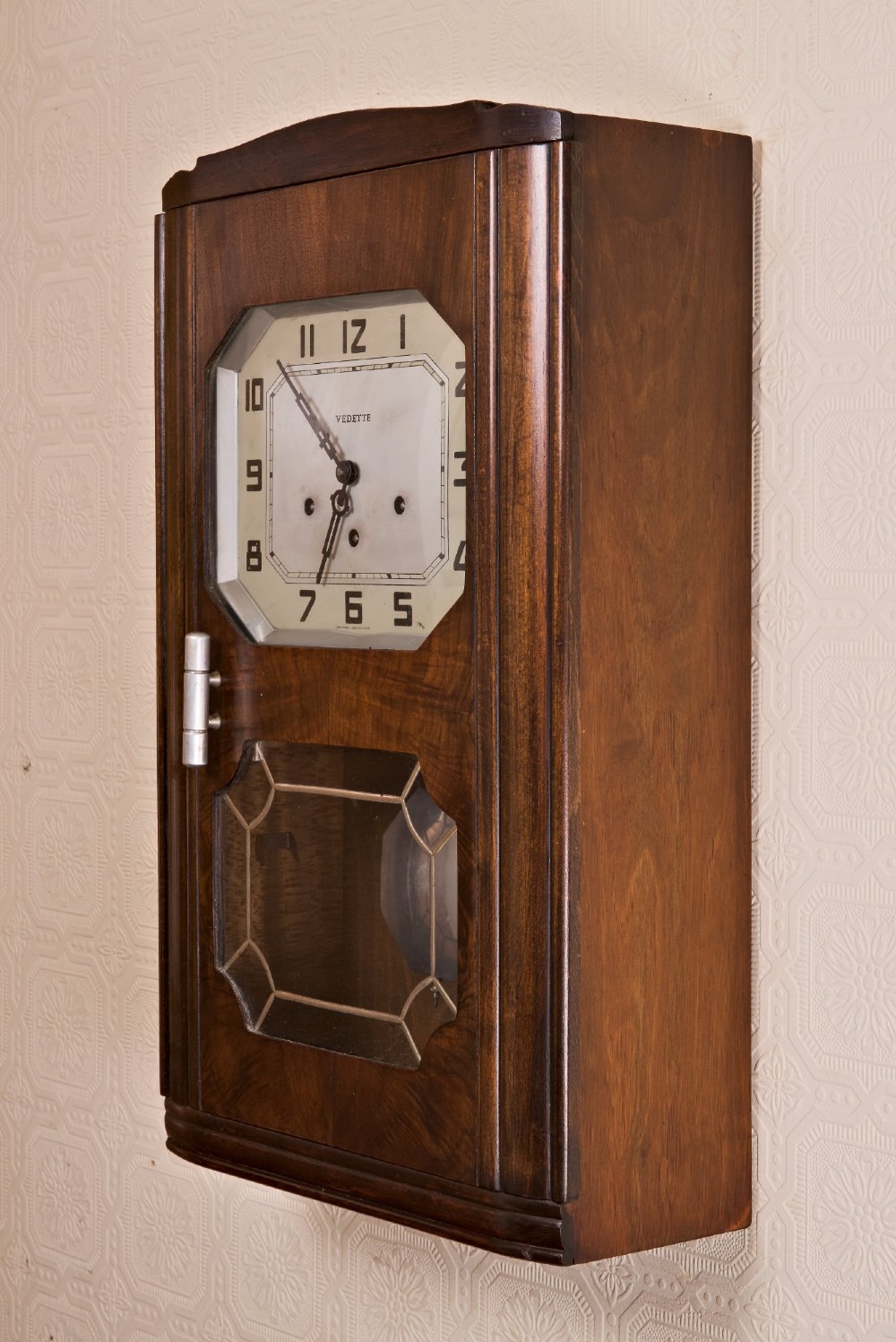 An Art Deco rectangular wooden wall clock featuring a light octagonal dial with Arabic numerals above a geometric leaded glass window. View VII