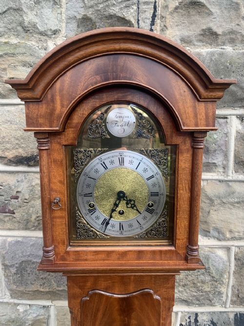 fig 1. A slim mahogany granddaughter clock featuring an arched top hood, brass and silvered dial, flame mahogany veneered trunk door, and bracket feet.