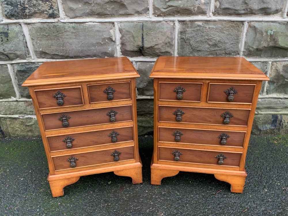 A matching pair of warm brown yewwood bedside chests featuring four drawer fronts with ornamental brass drop handles and bracket feet.