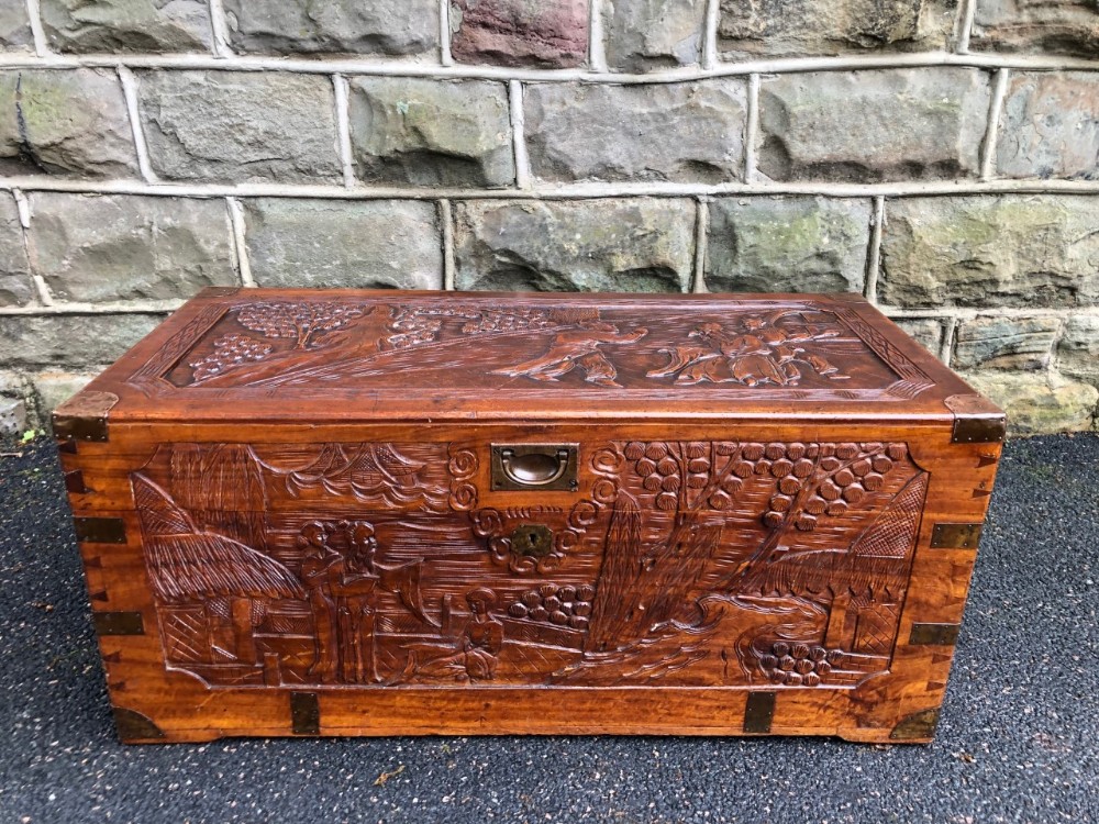 A rectangular carved camphor wood chest featuring relief scenes of figures and oriental architecture with brass corner brackets.