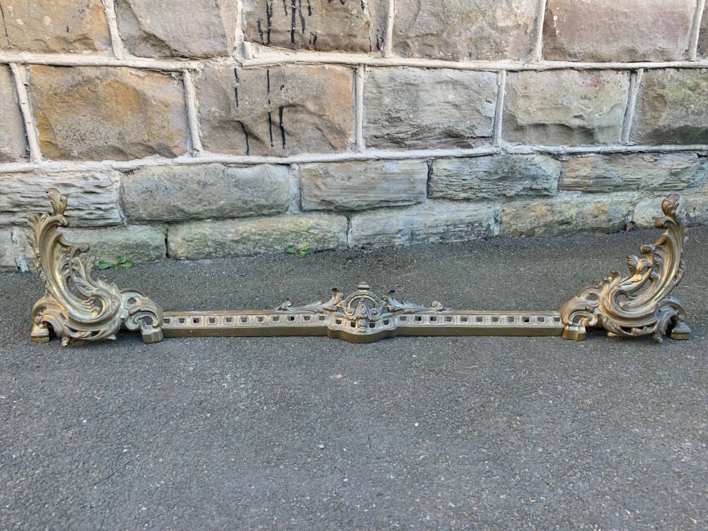 An adjustable cast brass fire fender featuring Rococo acanthus leaf scroll supports on both ends and a pierced central rail.