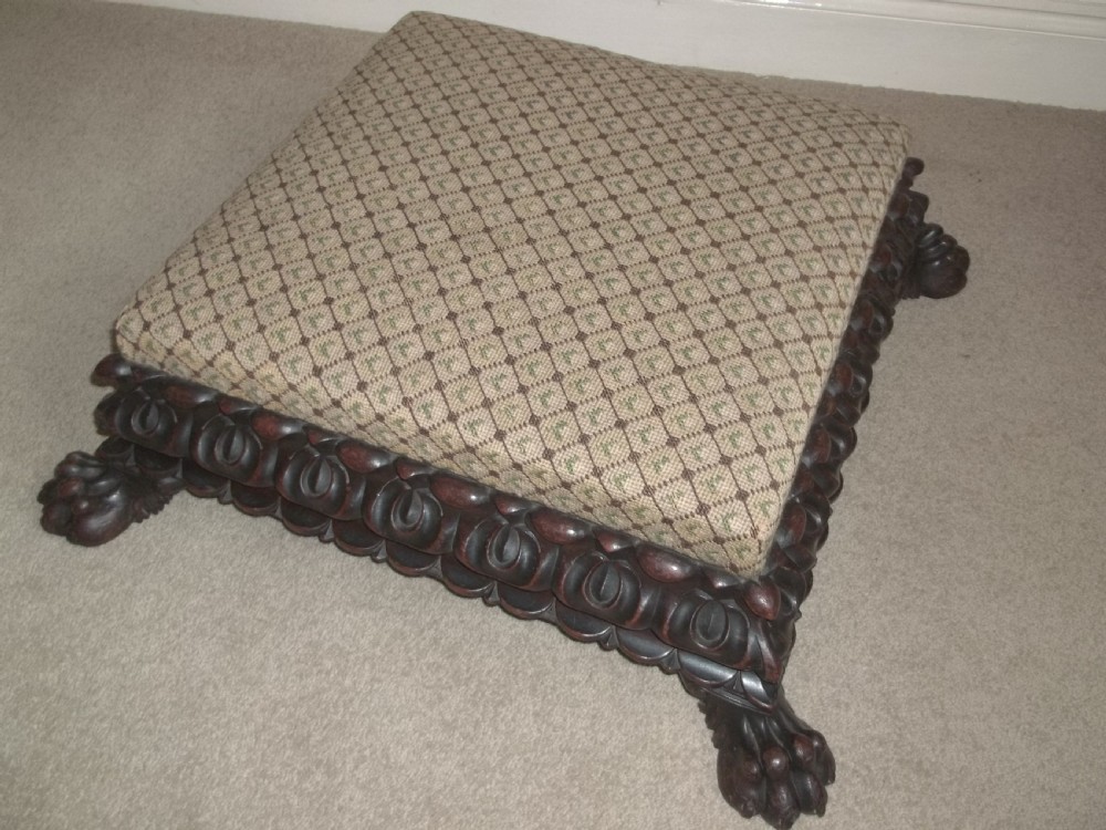 A low square William IV antique footstool with a deeply carved dark timber border, paw feet, and a beige patterned upholstered top.