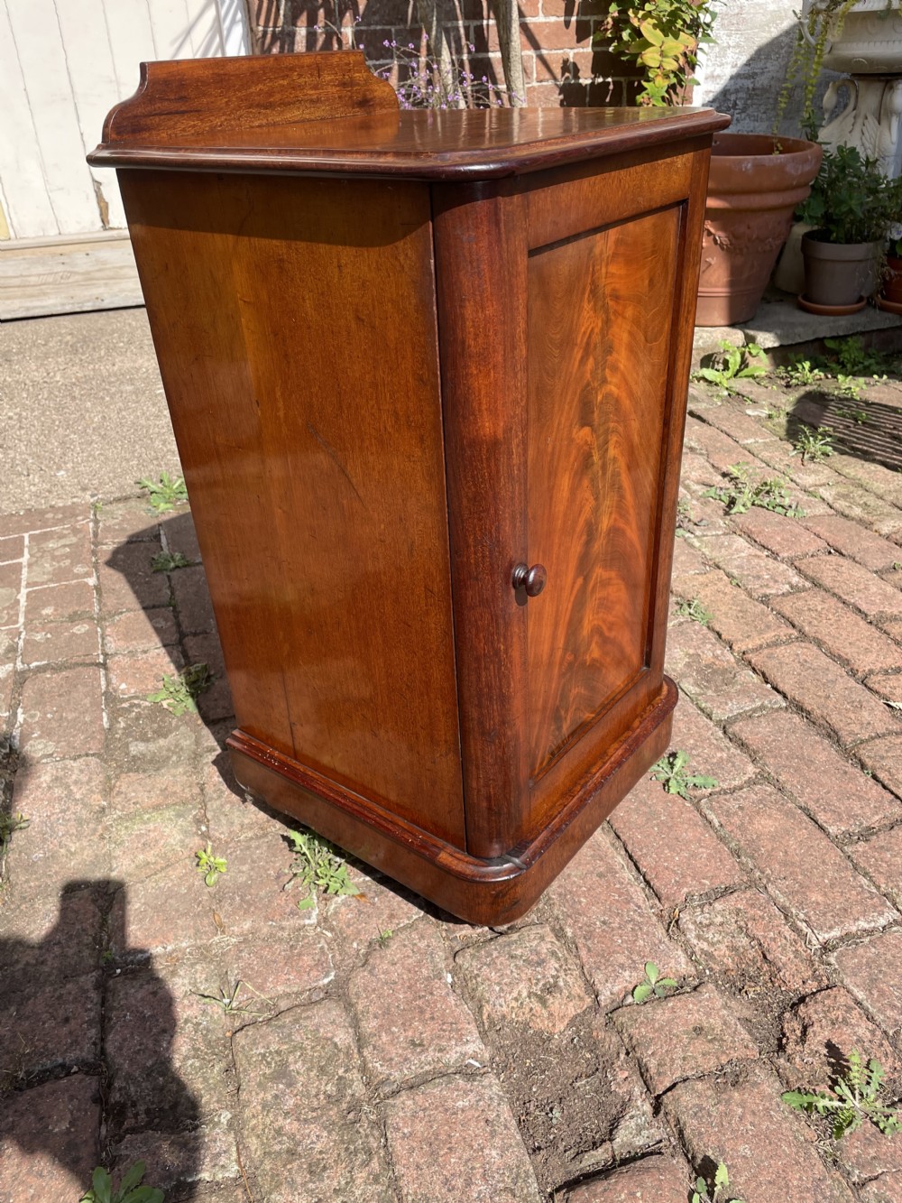 A Victorian mahogany bedside cabinet featuring a single panelled door with flame mahogany grain, a small turned knob, a plinth base, and a low gallery upstand. View VII
