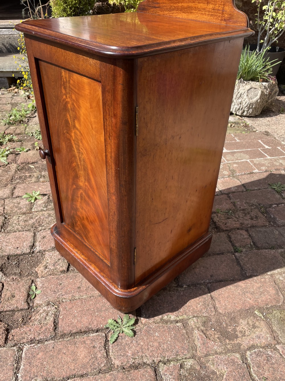 A Victorian mahogany bedside cabinet featuring a single panelled door with flame mahogany grain, a small turned knob, a plinth base, and a low gallery upstand. View VIII